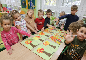 Dzieci stoją wokół stołu i prezentują swoje prace, które leżą na stole.