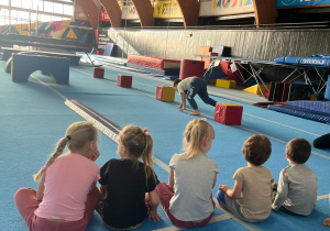 dzieci ćwiczą na niebieskiej macie na sali gimnastycznej, pokonują tor przeszkód