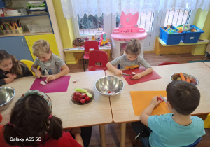 dzieci siedzą przy stolikach, kroją owoce i warzywa na kolorowych matach