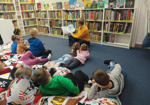 dzieci w bibliotece siedzą na dużych poduchach, słuchają opowiadania czytanego przez panią bibliotekarkę