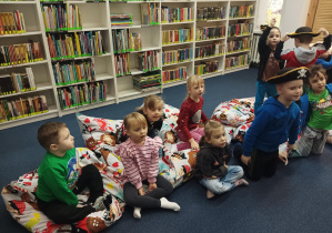 dzieci w bibliotece siedzą na dużych poduchach, słuchają opowiadania czytanego przez panią bibliotekarkę