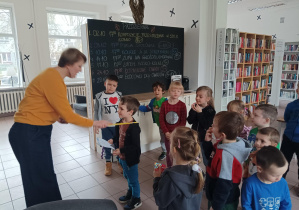 dzieci znajdują się w bibliotece, pani bibliotekarka wręcza dzieciom kartę biblioteczną