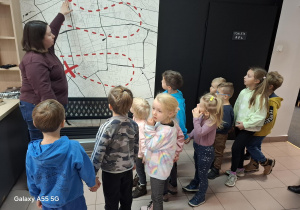 dzieci zwiedzają bibliotekę, pani bibliotekarka pokazuje palcem miejsce na mapie