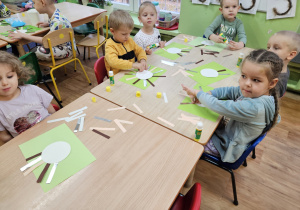 Dzieci siedzą przy stolikach i wykonują pracę plastyczną.