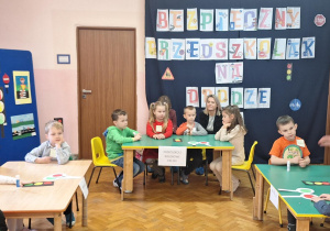 Dzieci siedzą przy stolikach i wykonują zadania.