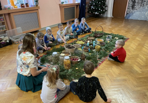 Dzieci siedzą wokół zgromadzonych ziół i naturalnych ozdób świątecznych.