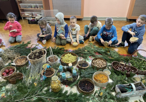 Dzieci siedzą wokół zgromadzonych ziół i naturalnych ozdób świątecznych.