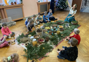 Dzieci siedzą wokół zgromadzonych ziół i naturalnych ozdób świątecznych.