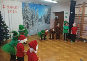 dzieci w przebraniach choinek, krasnoludków, zajączków występują na scenie, w tle zimowa dekoracja