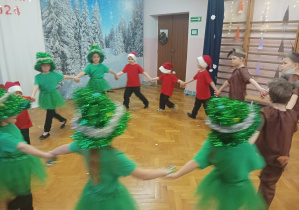 dzieci w przebraniach choinek, krasnoludków, zajączków występują na scenie, w tle zimowa dekoracja