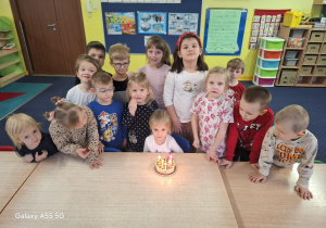 dziewczynka świętuje urodziny, przed nią na stoliku widać zabawkowy urodzinowy tort ze świeczką, za nią stoją dzieci