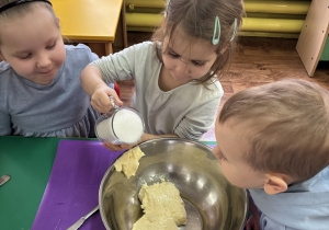 Dzieci siedzą przy stole, dziewczynka wsypuje kolejny produkt do miski.