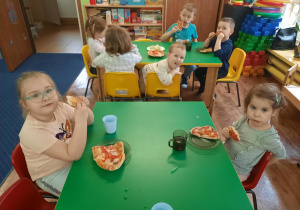 Dzieci siedzą przy stole i jedzą pizzę.