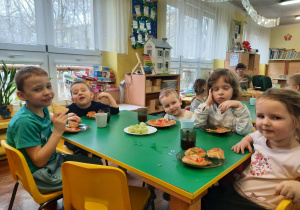 Dzieci siedzą przy stole i jedzą pizzę.