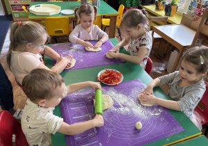 Dzieci siedzą przy stole i formują ciasto na pizzę.