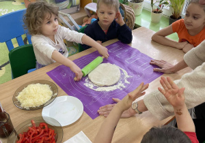 Dzieci siedzą przy stole, jedno z nich wałkuje ciasto na pizzę.