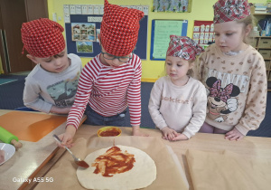 dzieci w kolorowych chustkach na głowie, przy stolikach, samodzielnie przygotowują pizzę.