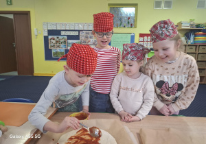 dzieci w kolorowych chustkach na głowie, przy stolikach, samodzielnie przygotowują pizzę.