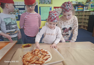 dzieci w kolorowych chustkach na głowie, przy stolikach, samodzielnie przygotowują pizzę.