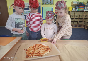 dzieci w kolorowych chustkach na głowie, przy stolikach, samodzielnie przygotowują pizzę.