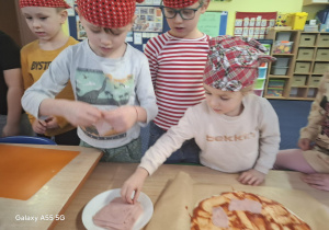 dzieci w kolorowych chustkach na głowie, przy stolikach, samodzielnie przygotowują pizzę.