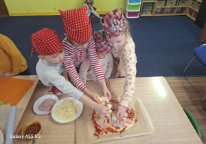 dzieci w kolorowych chustkach na głowie, przy stolikach, samodzielnie przygotowują pizzę.