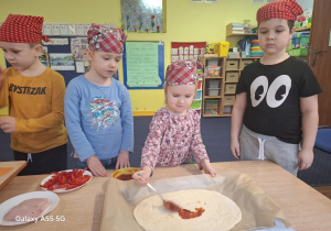 dzieci w kolorowych chustkach na głowie, przy stolikach, samodzielnie przygotowują pizzę.