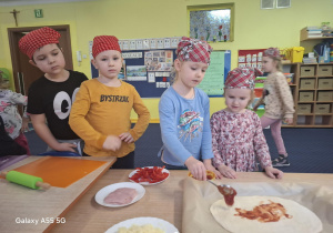 dzieci w kolorowych chustkach na głowie, przy stolikach, samodzielnie przygotowują pizzę.