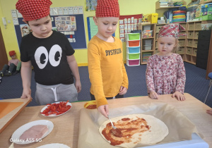 dzieci w kolorowych chustkach na głowie, przy stolikach, samodzielnie przygotowują pizzę.