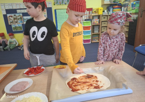 dzieci w kolorowych chustkach na głowie, przy stolikach, samodzielnie przygotowują pizzę.