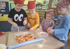 dzieci w kolorowych chustkach na głowie, przy stolikach, samodzielnie przygotowują pizzę.