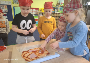 dzieci w kolorowych chustkach na głowie, przy stolikach, samodzielnie przygotowują pizzę.