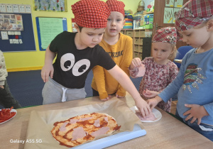 dzieci w kolorowych chustkach na głowie, przy stolikach, samodzielnie przygotowują pizzę.