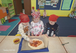 dzieci w kolorowych chustkach na głowie, przy stolikach, samodzielnie przygotowują pizzę.