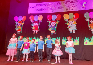 dzieci stoją na scenie Monopolis, śpiewają piosenkę, grają na instrumentach