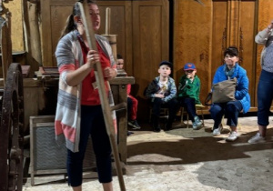 Dzieci siedzą w stodole i oglądają sprzęty wykorzystywane dawniej na wsi.
