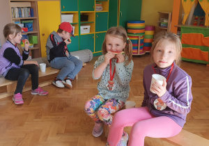 Dzieci siedzą na ławeczkach pijąc i zjadając owoce.