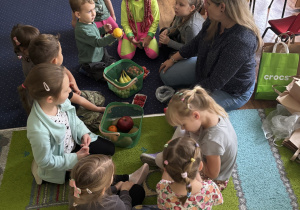 Dzieci siedzą na dywanie, oglądają i nazywają zakupione owoce.