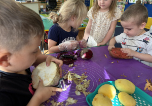 Dzieci siedzą przy stole i obierają warzywa.