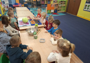 Dzieci siedzą przy stole na którym leżą warzywa i przedmioty niezbędne do kiszenia kapusty.