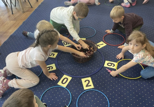 Dzieci siedzą na dywanie i rozwiązują zadania matematyczne.