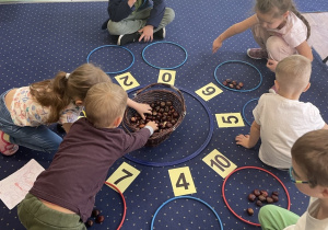 Dzieci siedzą na dywanie i rozwiązują zadania matematyczne.