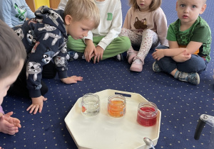 Dzieci siedzą w kole na dywanie i obserwują słoiki.