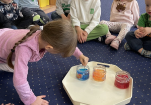 Dzieci siedzą na dywanie, dziewczynka miesza wodę łyżeczką.