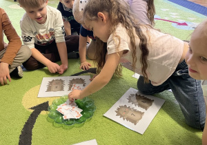 Dzieci siedzą na dywanie w kole i wykonują zadanie.