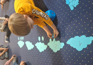 Dzieci siedzą na dywanie, do chmurek mocują odpowiednią liczbę kropel.