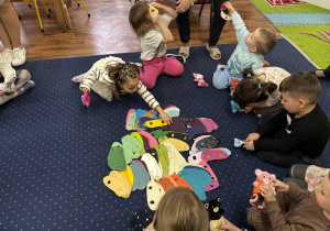 Dzieci siedzą na dywanie i wybierają kamizelki dla swojego misia.
