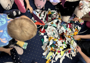 Dzieci siedzą na dywanie i wybierają apaszki dla swojego misia.