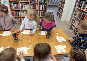 Dzieci stoją przy stole i ozdabiają świąteczne kartki.