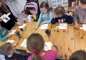 Dzieci stoją przy stole i ozdabiają świąteczne kartki.